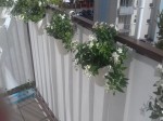 Biała osłona balkonowa na wymiar zamontowana na balustradzie tarasu, ozdobiona wiszącymi doniczkami z kwiatami.