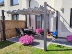 Szara roleta rzymska pozioma premium Decor 2,5x4 zamontowana na grafitowej pergoli nad tarasem z meblami i huśtawkami.