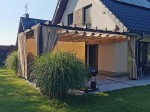 Roleta rzymska pozioma premium Decor 4x4 na ciemnej konstrukcji pergoli przy domu z jasną elewacją i tarasem.