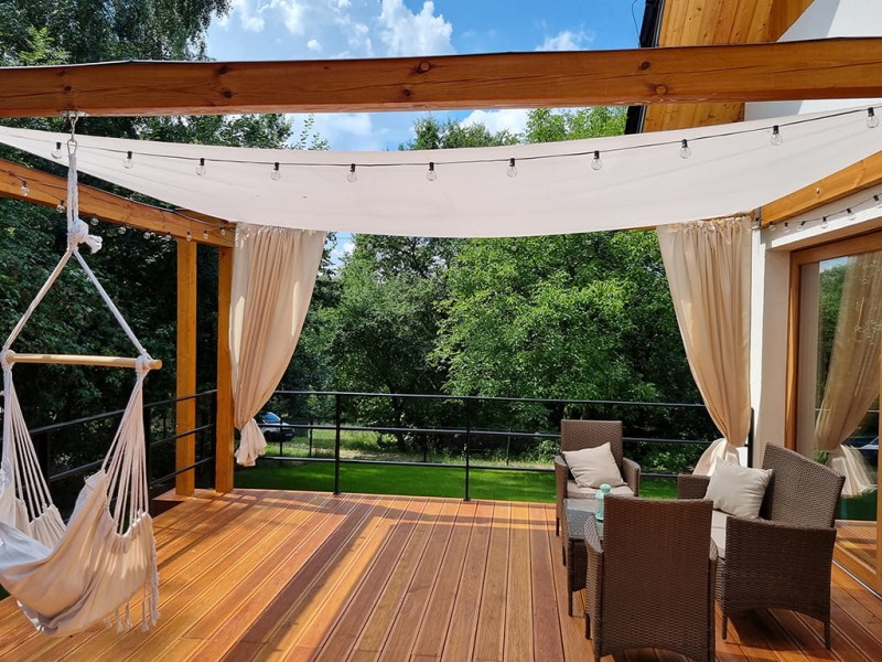 Biały żagiel przeciwsłoneczny 3,2x4,2 m zamontowany nad tarasem z wypoczynkiem i wiszącym fotelem.
