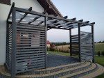Biały pas altanowy przeciwsłoneczny 1x5 m przeplatany przez belki grafitowej pergoli nad tarasem z kostki brukowej.