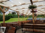 Biały pas altanowy przeciwsłoneczny na wymiar zamontowany na drewnianej konstrukcji pergoli w ogródku restauracyjnym.