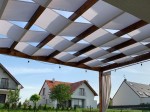 Biały wodoszczelny pas altanowy 0,5x5 m przeplatany przez drewniane belki pergoli ogrodowej na tle domów jednorodzinnych.