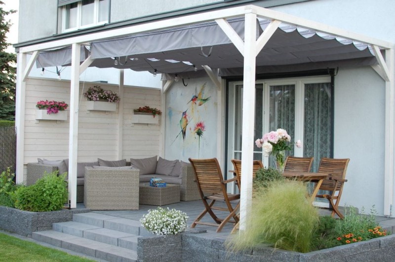 Popielata pozioma roleta rzymska o wymiarach 2,68x2,13 m zamontowana na białej pergoli nad tarasem z meblami.