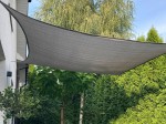 Szary żagiel przeciwsłoneczny HDPE 5x6 rozwieszony na tarasie przy domu, otoczony gęstą, zieloną roślinnością.