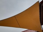 Żółty trójkątny żagiel przeciwsłoneczny przemysłowy wykonany z wodoszczelnej tkaniny, zamontowany nad tarasem.