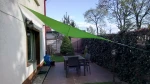 Zielony trójkątny żagiel ogrodowy zamontowany nad tarasem z zestawem mebli ogrodowych i roślinnością w tle.