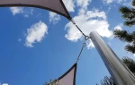 Beżowy żagiel przeciwsłoneczny wodoszczelny zamontowany za pomocą metalowych uchwytów do stalowego masztu na tle nieba.