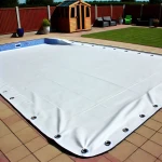 Biała plandeka na basen na wymiar z oczkami montażowymi, przykrywająca przydomowy basen w ogrodzie.