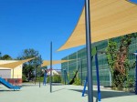 Trójkątny żagiel przeciwsłoneczny HDPE 3x3x4,2 m w kolorze beżowym zamontowany nad nowoczesnym placem zabaw.