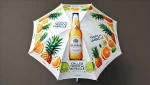 Biały parasol ogrodowy z grafiką butelki napoju Deskea, plastrami cytrusów i ananasami w tropikalnym stylu.