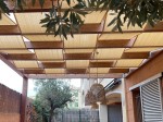 Beżowy pas altanowy przeciwsłoneczny HDPE zamontowany na drewnianej pergoli nad tarasem z lampami i drzewem.
