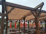Beżowa pozioma roleta rzymska zamontowana na drewnianej pergoli w ogródku restauracyjnym z drewnianymi meblami.