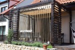 Beżowa roleta rzymska pozioma zamontowana na drewnianej altanie z ozdobnymi sznurami i meblami ogrodowymi.