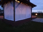 Biała zasłona firankowa na wymiar zamontowana w drewnianej altanie ogrodowej o zmierzchu.