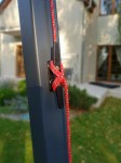 Plastikowa czarna knaga rogowa zamontowana na szarym słupku, zabezpieczająca czerwoną lino do montażu żagli przeciwsłonecznych.