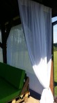 Biała zasłona meshflag o wymiarach 1,29x2,24 m zamocowana w drewnianej altanie obok zielonej sofy ogrodowej.