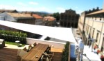 Biały trójkątny żagiel przeciwsłoneczny 3,2x3,2x4,5 m zamontowany nad tarasem z drewnianym stołem i meblami.