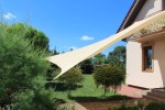 Beżowy żagiel przeciwsłoneczny HDPE o wymiarach 5x6 metrów zamontowany nad tarasem obok domu jednorodzinnego.