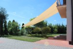 Beżowy żagiel przeciwsłoneczny HDPE 5x5 m zamontowany na tarasie nad ogrodem zapewnia cień w słoneczny dzień.