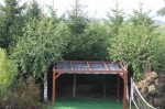 Brązowa pergola z zamontowanym szarym żaglem przeciwsłonecznym rozsuwanym HDPE w otoczeniu gęstych drzew.