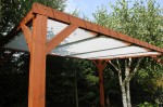 Brązowa drewniana pergola z zamontowanym białym żaglem przeciwsłonecznym typu mesh na wymiar w ogrodzie.