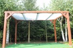 Brązowa drewniana pergola z dachem żaglowym z białej siatki mesh w zielonym ogrodzie.