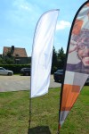 Biała flaga reklamowa typu husarz na pałąku ustawiona na trawniku przed budynkiem, obok zadrukowanego żagla.
