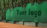Zielona osłona na korty tenisowe na wymiar z logo, zamontowana na ogrodzeniu przy ceglanej mączce i zieleni.
