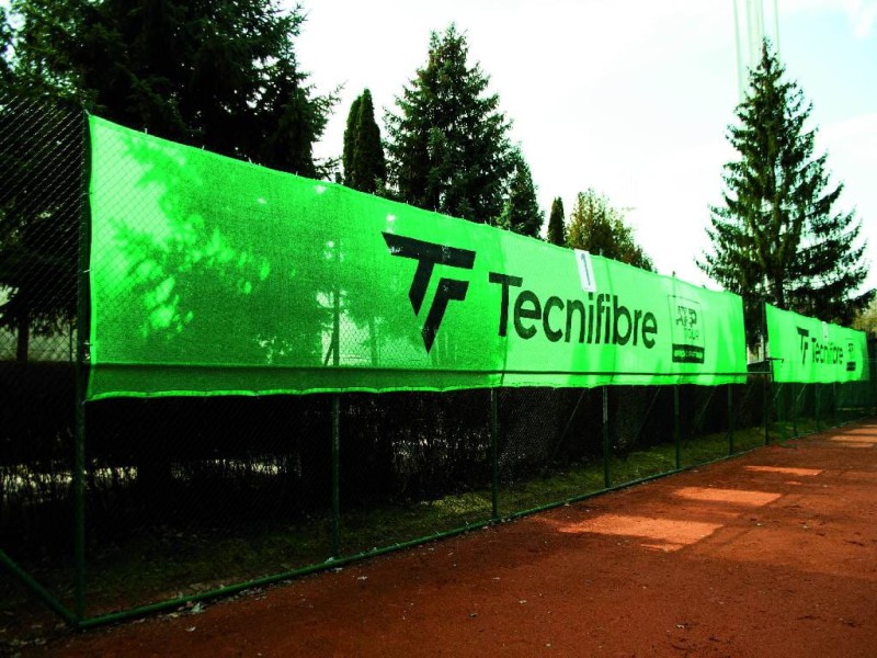 Zielona osłona na korty tenisowe na wymiar z nadrukiem marki Tecnifibre zamontowana na ogrodzeniu kortu ziemnego.
